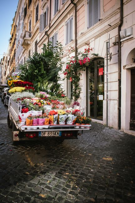 A delivery truck parked on a cobblestone street in front of a building with cream-colored walls and shuttered windows, loaded with a variety of fresh flowers and floral arrangements. The flowers inclu