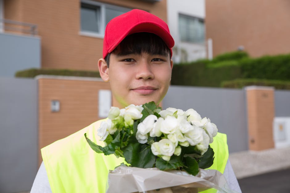A young male flower delivery courier wearing a red cap and a neon yellow vest is standing outdoors in front of modern residential buildings. He is holding a bouquet of fresh white flowers, likely rose